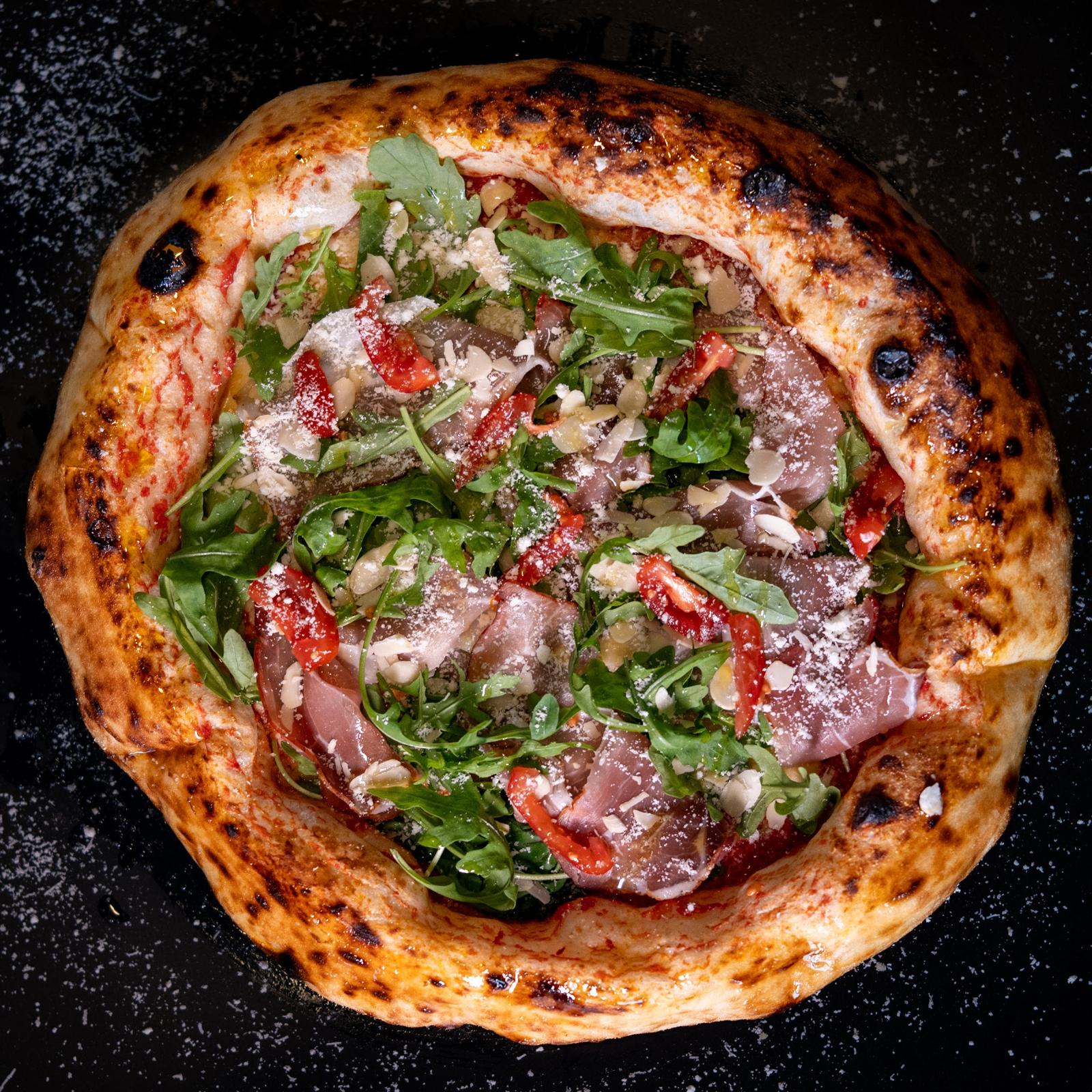 Pizza napoletană cu prosciutto crudo, rucola și Grana Padano, pe fundal negru presărat cu făină